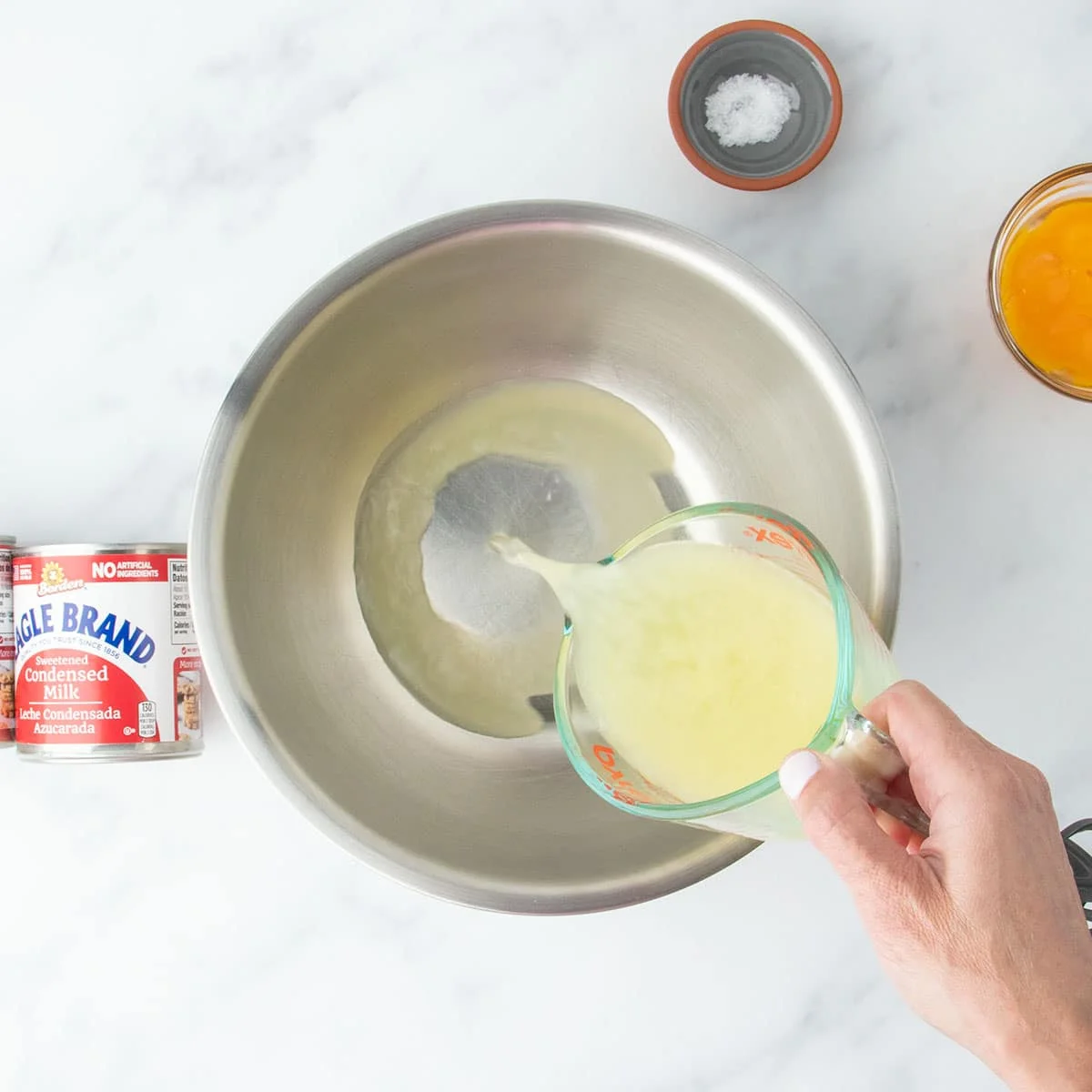 Lemon juice being poured into a large mixing bowl, surrounded by other ingredients on a marble surface.