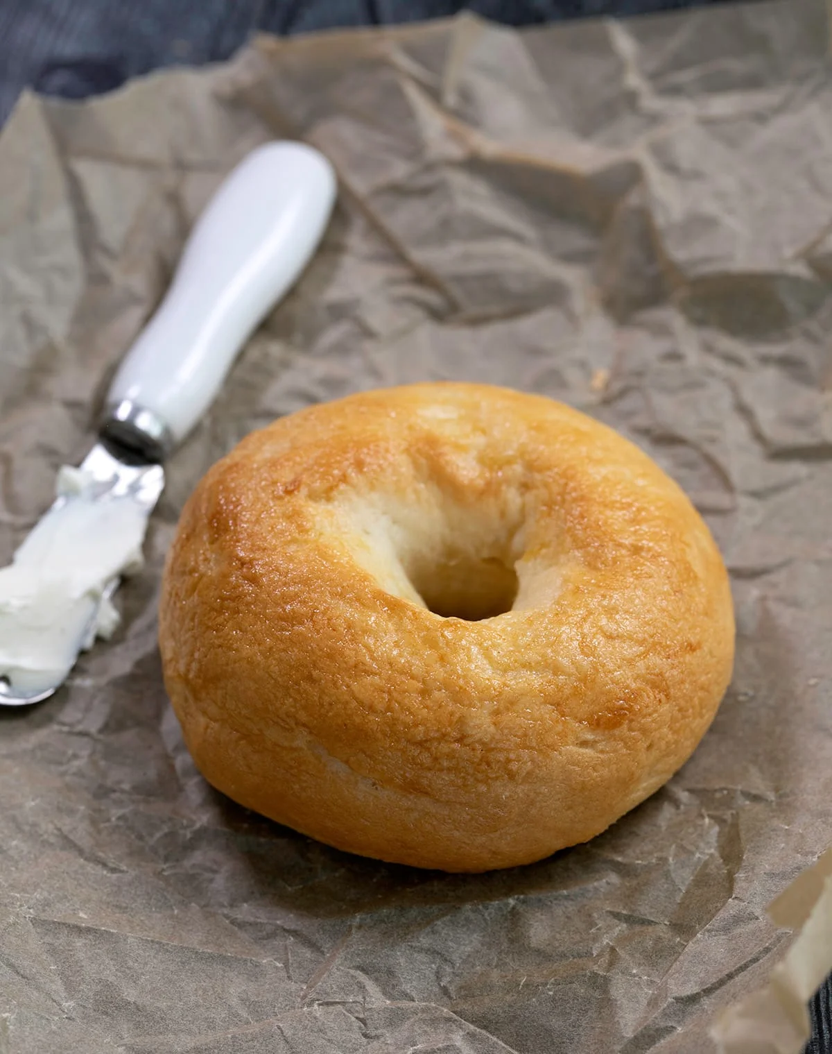 Light brown bagel on brown paper with a knife and cream cheese on a dish