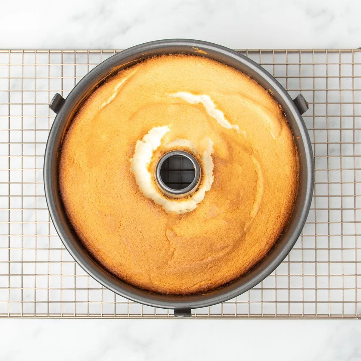 light brown baked angel food cake in tube pan on gold colored wire rack