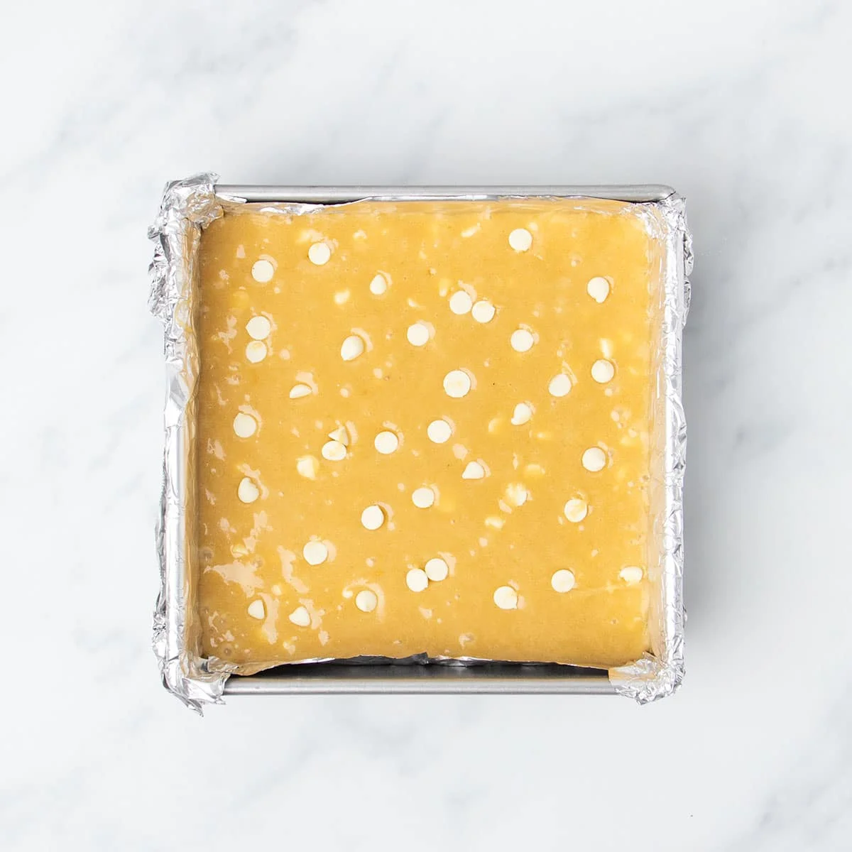 light brown raw blondie batter with white chocolate chips on top in foil-lined square metal baking pan