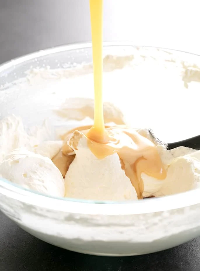 Light brown sweetened condensed milk being poured into glass bowl with white fluffy whipped cream