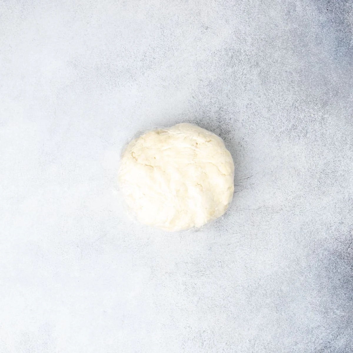 Light tan disk of pie crust wrapped in plastic wrap on light gray surface.