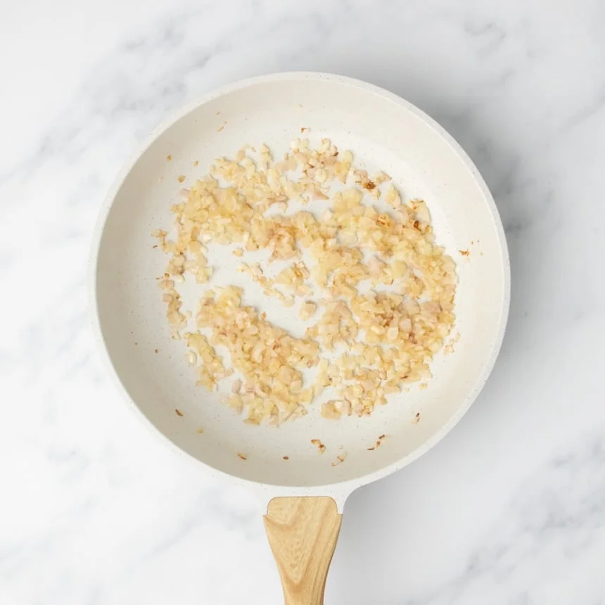 Lightly browned chopped onions cooking in a white skillet on a marble surface.