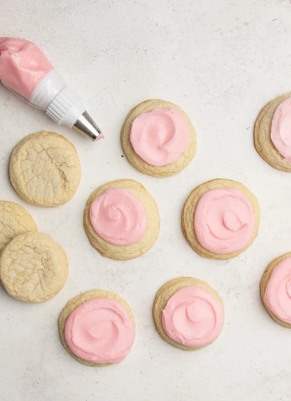 many light yellow sugar cookies on a white background, many with a swirl of pink frosting on top