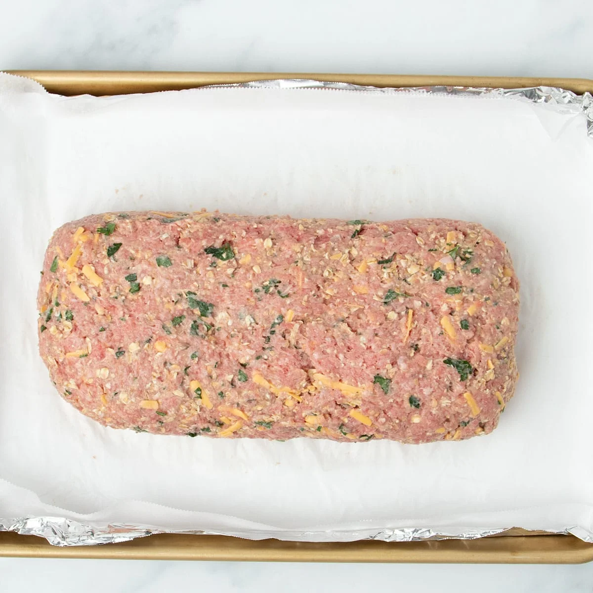 meatloaf mixture shaped into 10-inch x 5-inch rectangular loaf on white paper on top of aluminum foil on gold rimmed baking pan