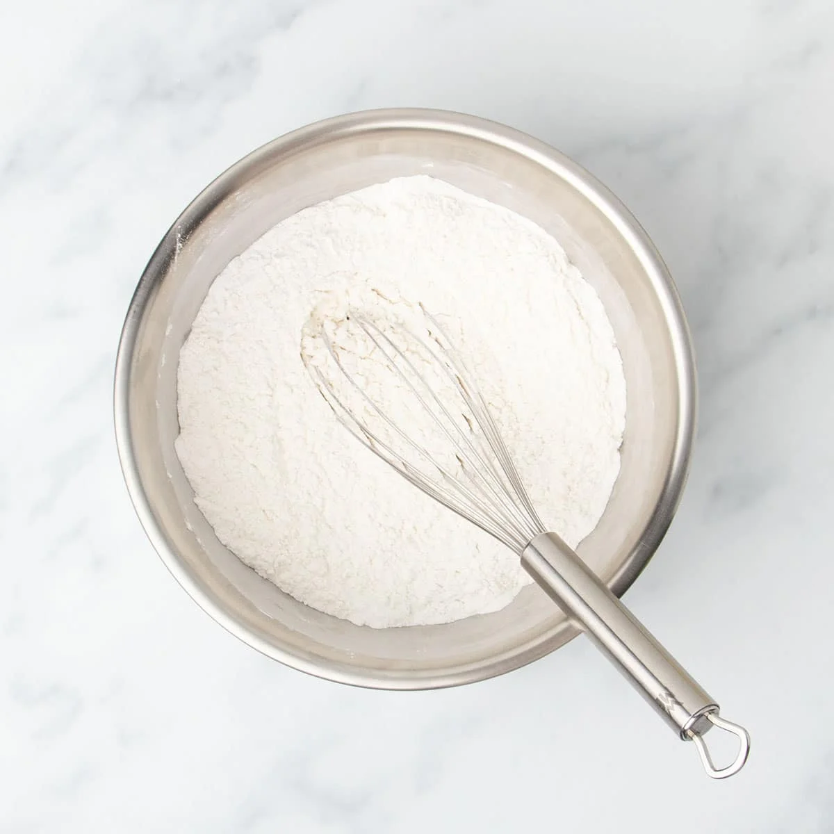 medium round metal mixing bowl with white flour and large metal wire whisk on marble surface