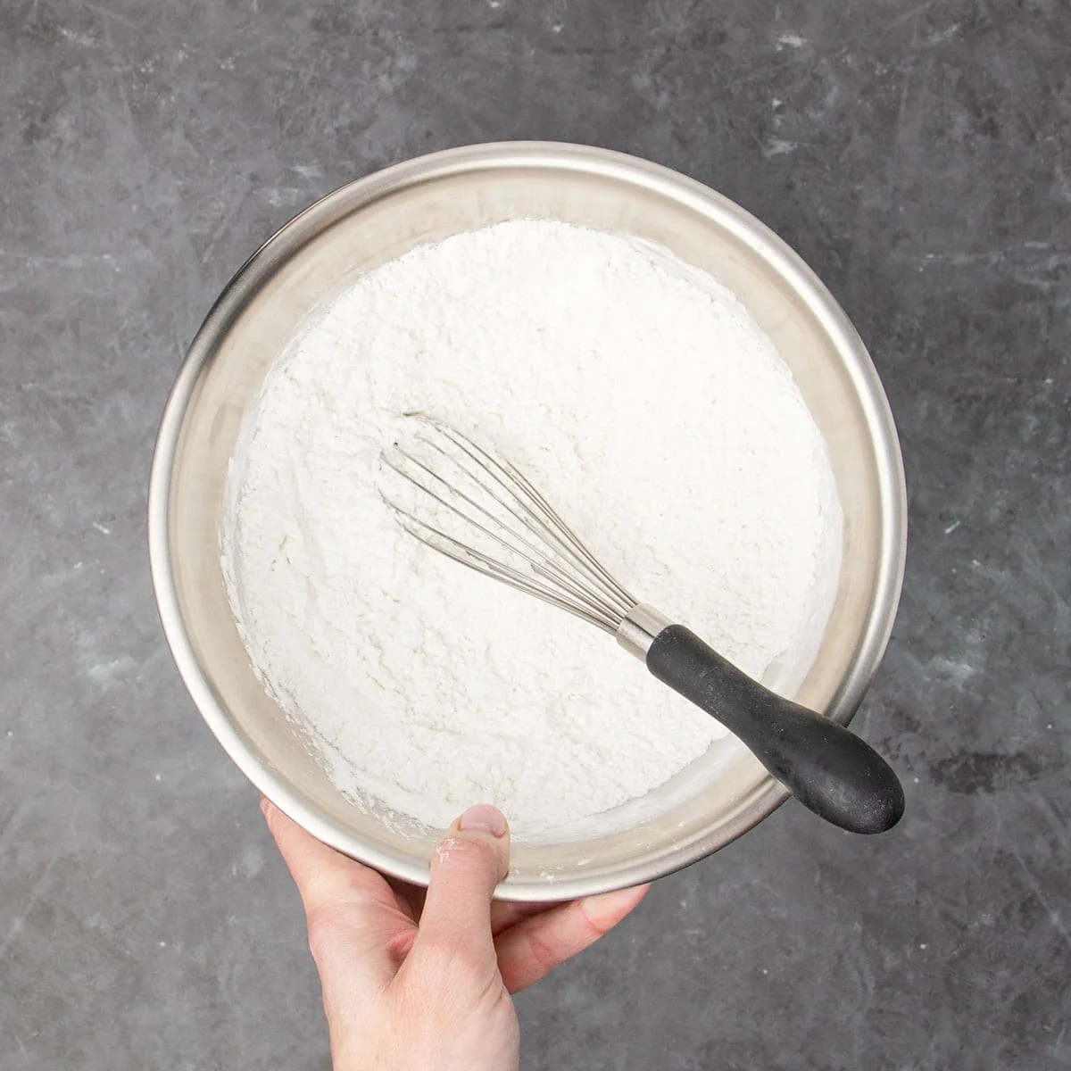 medium size round metal mixing bowl with white flour and wire whisk with black handle all being held in a hand