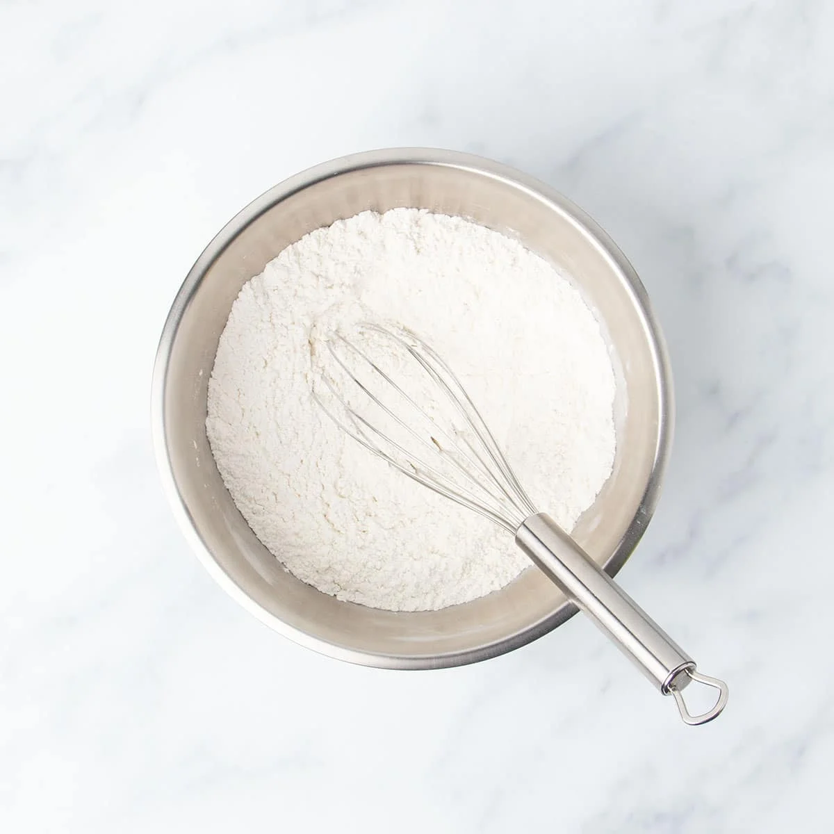 medium-size round metal mixing bowl with white flour powder and large wire whisk