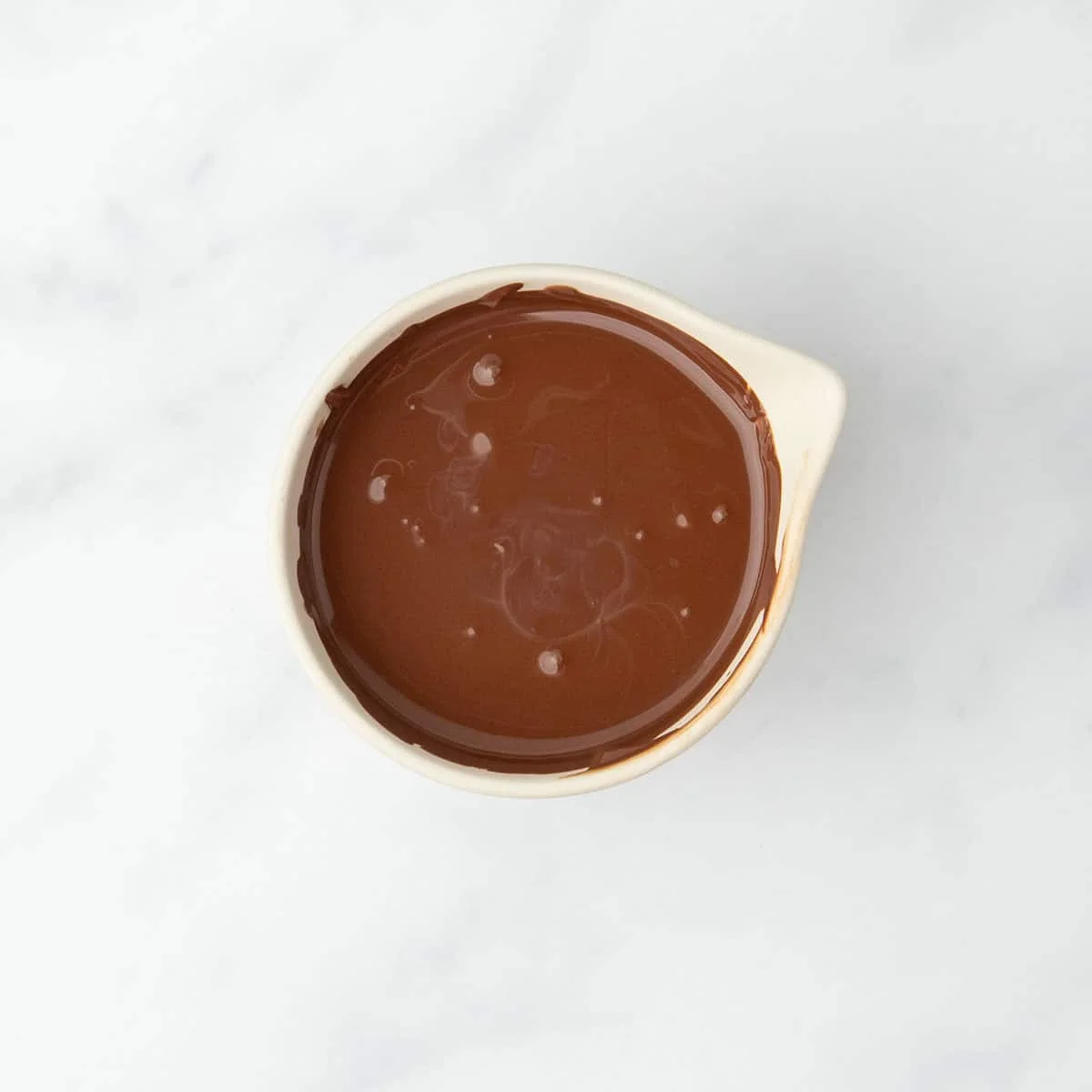 Melted dark chocolate swirled smooth in a white spouted bowl on a marble surface