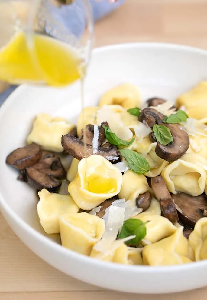 Melted garlic butter pouring onto tortellini with cooked mushrooms in white bowl.