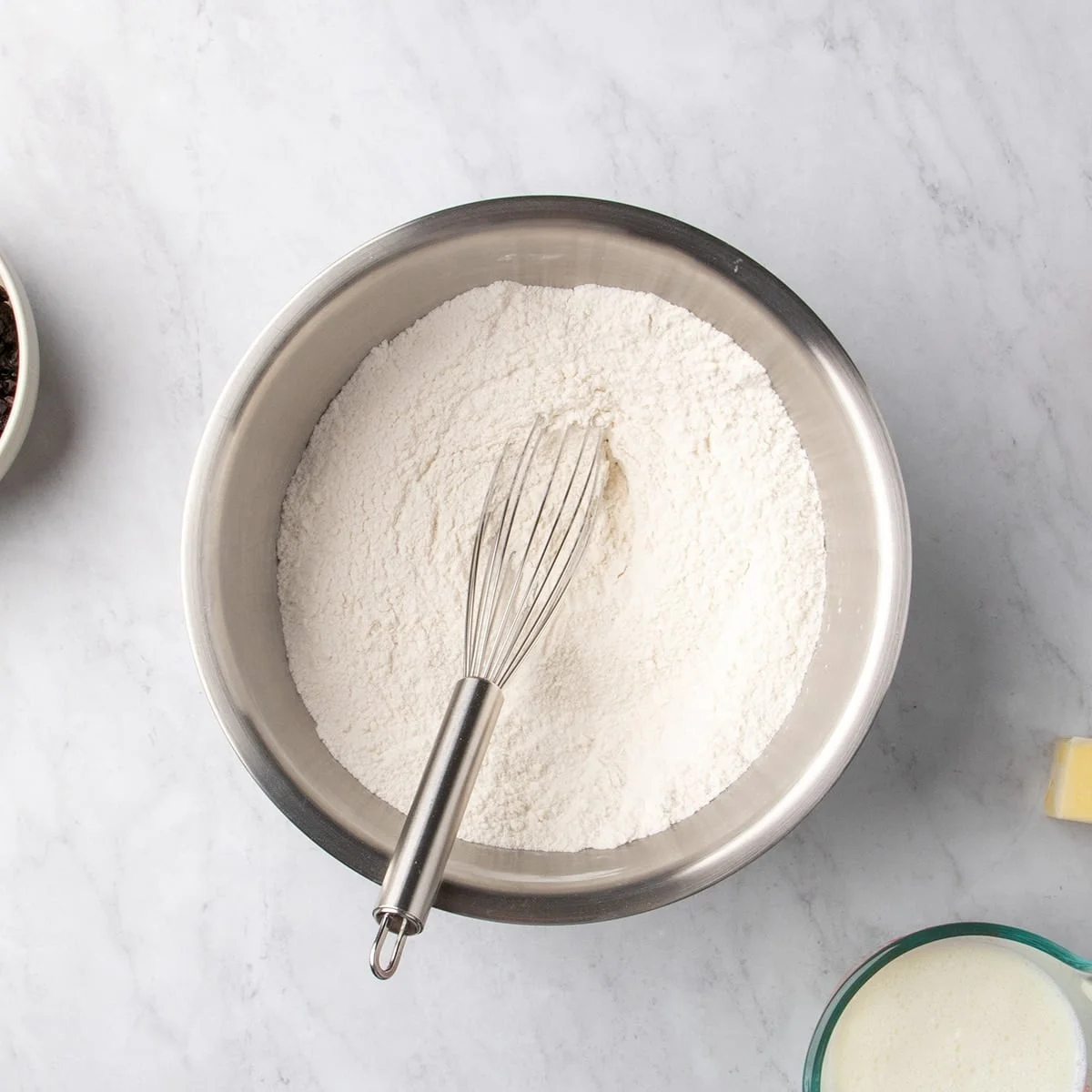 Metal mixing bowl filled with white dry ingredients and a metal whisk resting in the center on a marble surface.