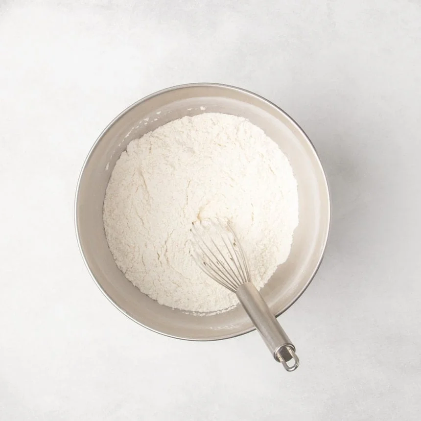 Metal mixing bowl with dry ingredients blended together and a whisk resting inside the bowl.