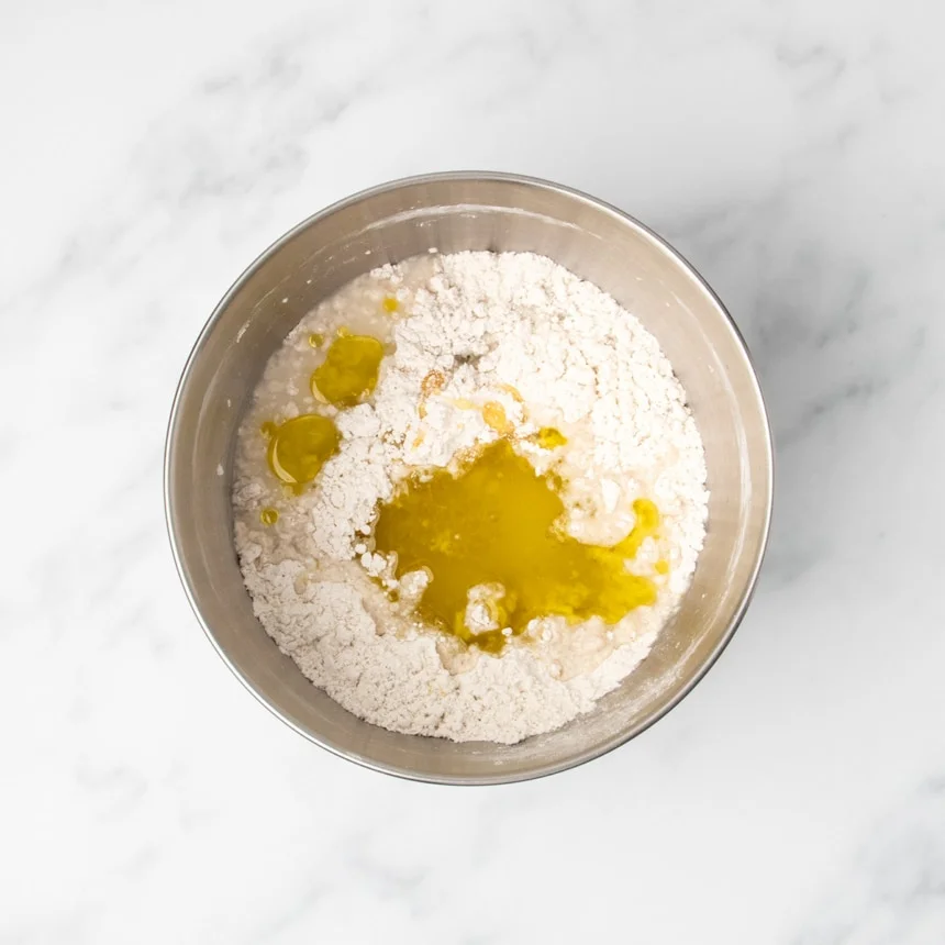 Metal round mixing bowl with dry flour mixture with olive oil, honey, water and vinegar added but not mixed in.