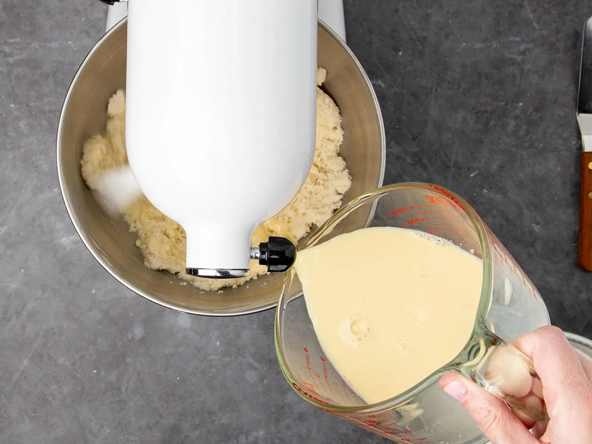 mixer on low speed while hand holding measuring cup pours yellow liquid into mixing bowl