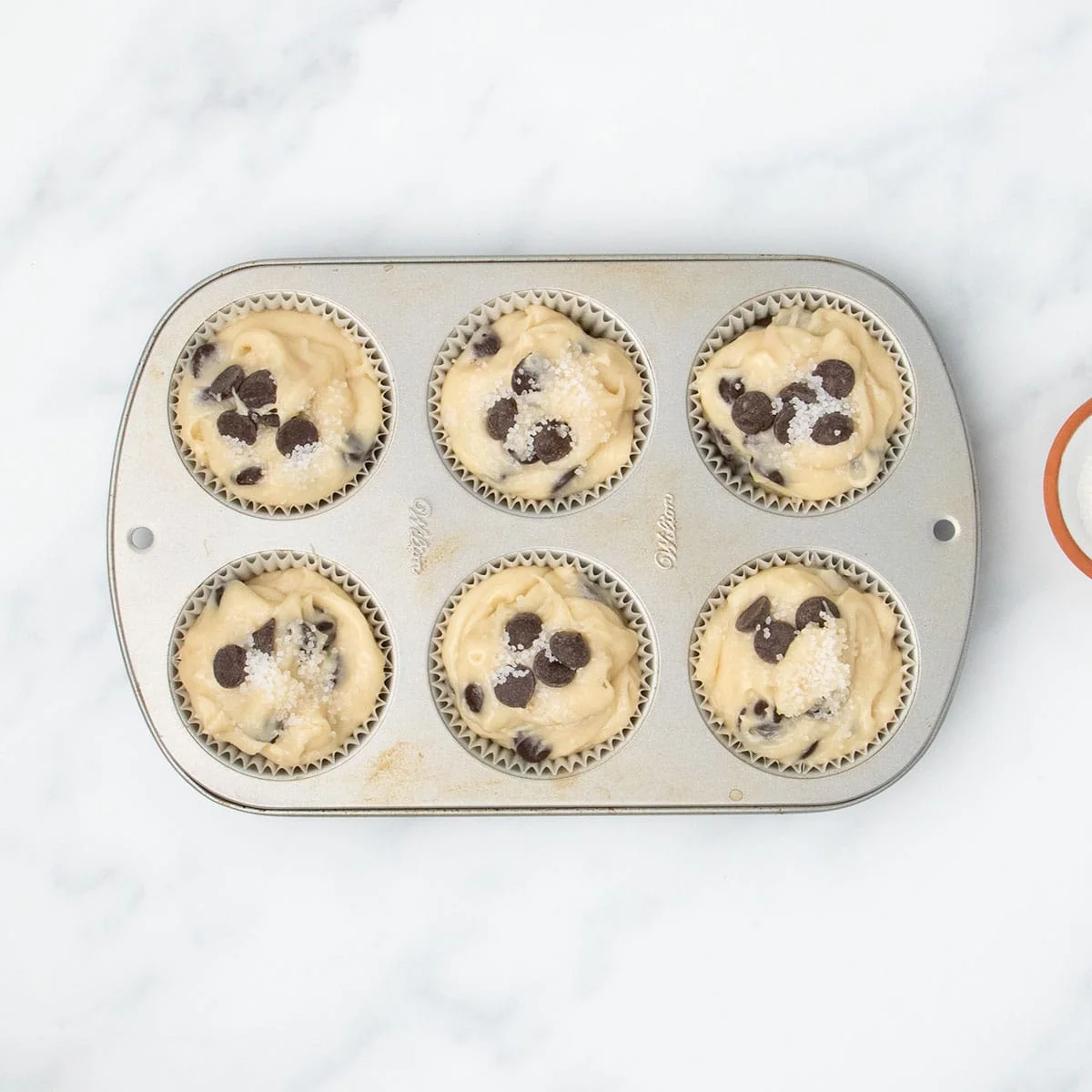Muffin batter portioned into a 6-cup muffin tin lined with muffin liners, topped with extra chocolate chips and coarse sugar.