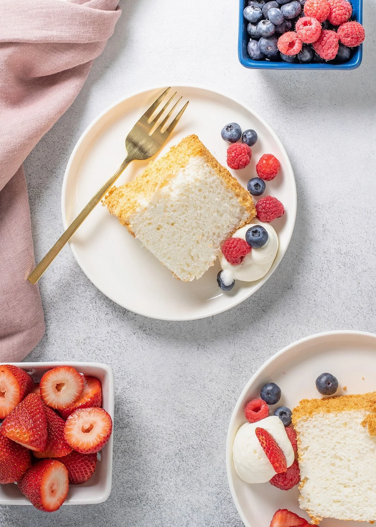 Overhead image of 2 white plates with gluten free angel food cake slice berries and cream one with a fork and extra berries