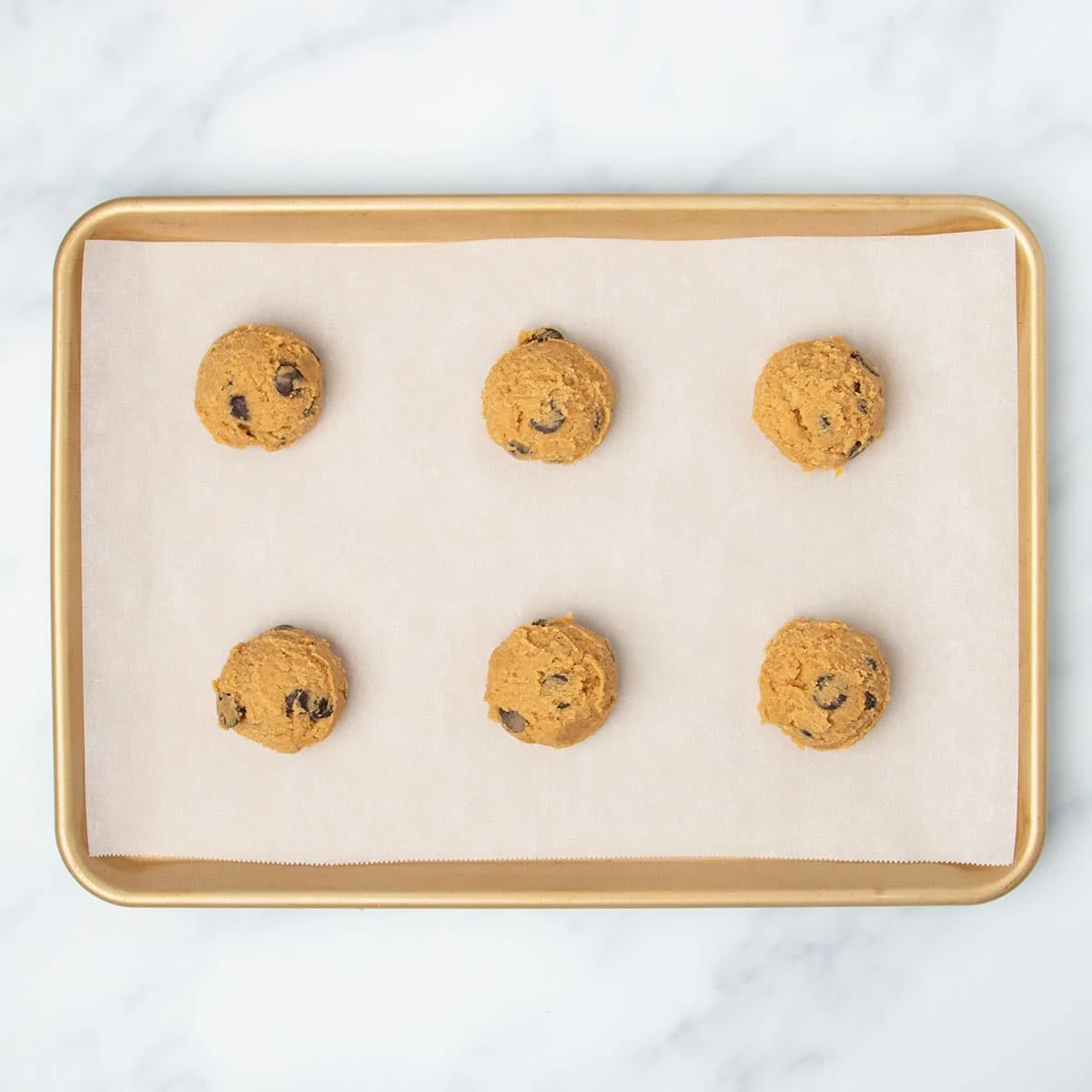 Overhead image of 6 raw rounds of light orange cookie dough with chocolate chips spaced evenly apart on white paper on gold rimmed baking sheet