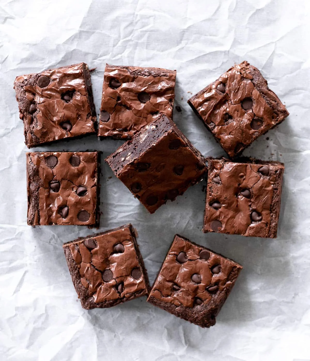 Overhead image of 8 brown bar squares with chocolate chips on white crinkly paper.