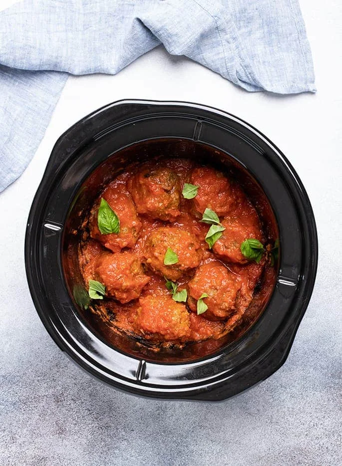 Overhead image of 8 cooked meatballs in tomato sauce in black crock pot liner with fresh basil pieces scattered on top
