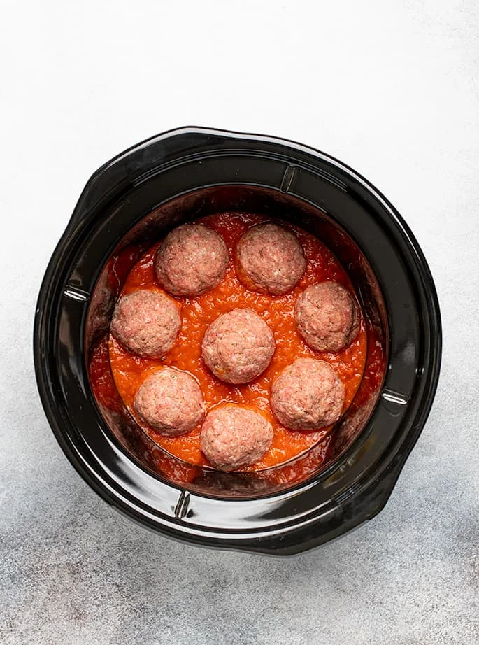 Overhead image of 8 raw meatballs in tomato sauce in round black crock pot liner
