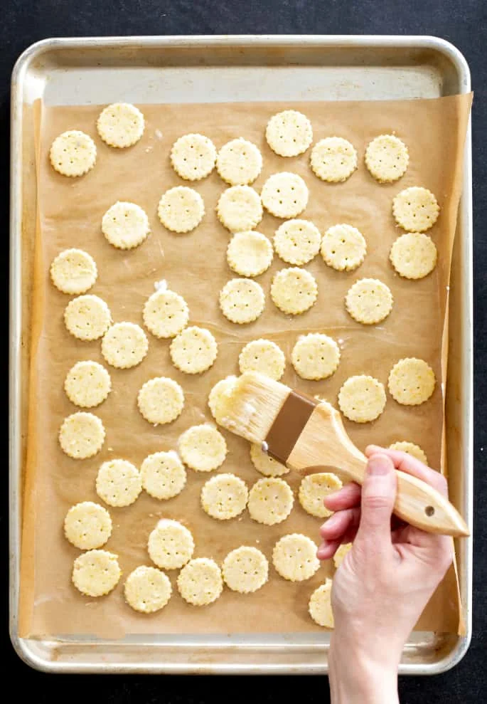 Overhead image of a hand holding a pastry brush and brushing raw cracker dough.