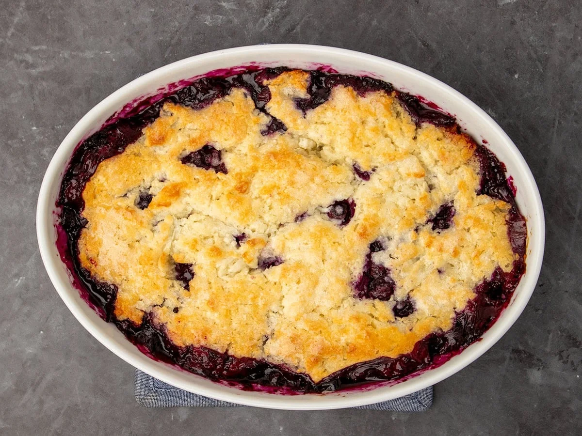 overhead image of baked bubbling blueberry cobbler filling with baked golden brown pastry topping in white casserole dish
