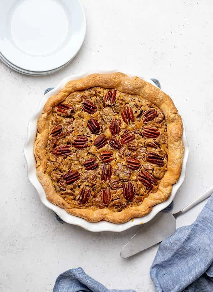 Overhead image of baked gluten free pecan pie with metal pie server, stack of small white round plates and blue cloth on white surface