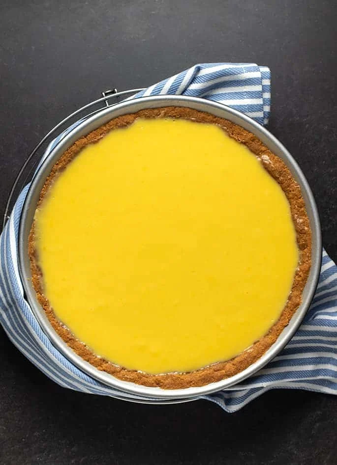 Overhead image of baked key lime pie filling in graham cracker crust in metal pie plate with blue and white striped towel on top of round wire rack