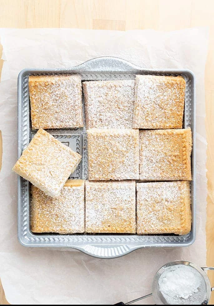 Overhead image of cut shortbread squares in pan with powdered sugar on white paper