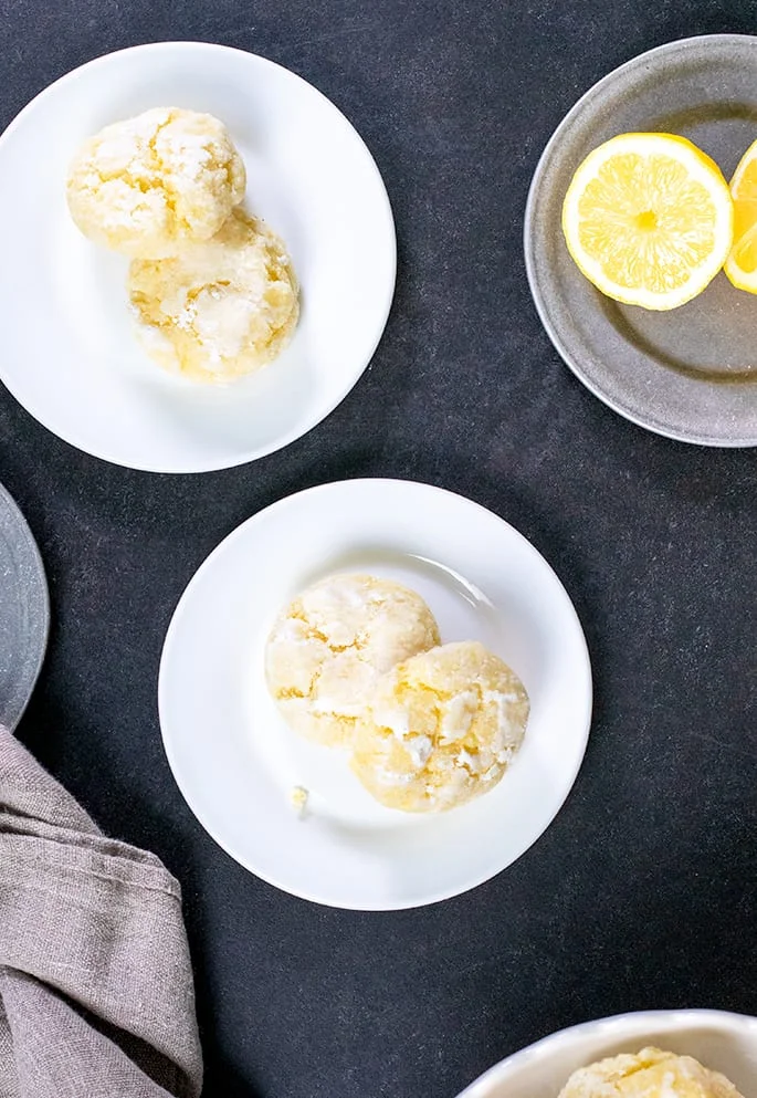 Overhead image of four lemon crinkle cookies on two small white plates and a cut lemon on a small metal plate