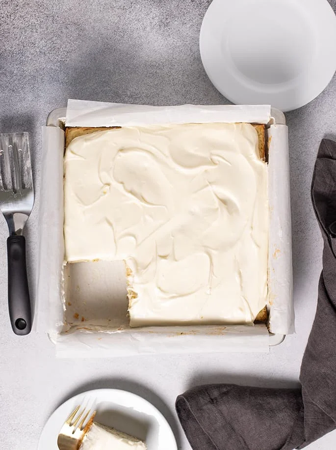 Overhead image of frosted zucchini cake in square pan with two small white plates and dark gray cloth