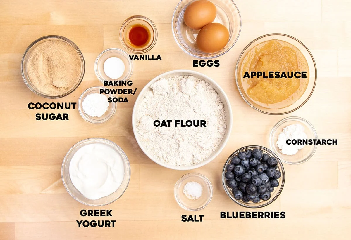 overhead image of ingredients for healthy blueberry muffins in small bowls with names of ingredients in bold black type on blond wood surface