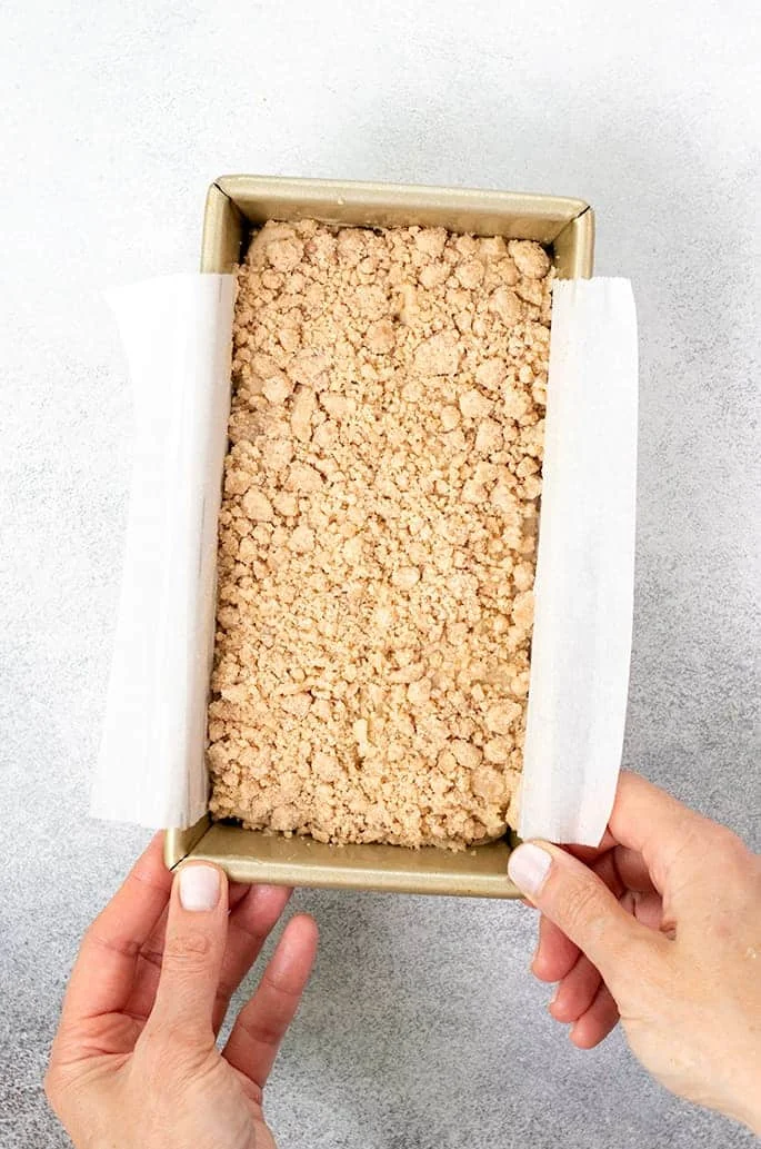 Overhead image of raw apple butter bread batter with crumble topping in loaf pan with hands on corners