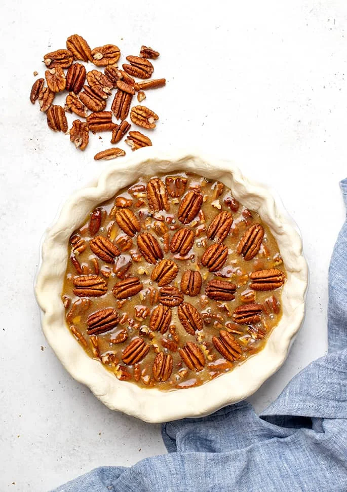 Overhead image of raw gluten free pecan pie with extra pecans on white surface and blue cloth