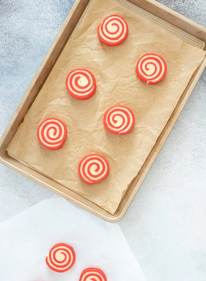 Overhead image of raw red and white pinwheel cookies on white paper and on lined baking tray.