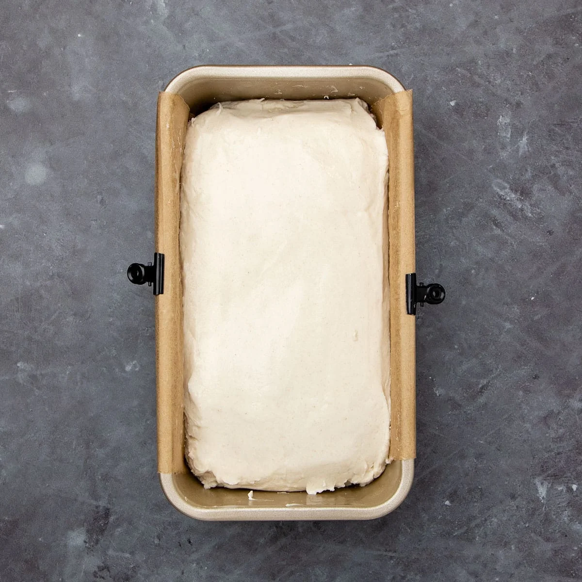 Overhead image of raw unrisen sourdough bread dough in single smooth layer in gold standard loaf pan lined with brown parchment paper.