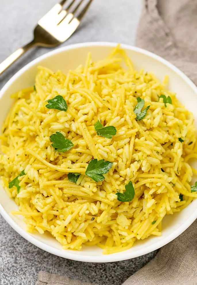 Overhead image of white round serving bowl with rice a roni with fresh parsley pieces, tan cloth, and fork.