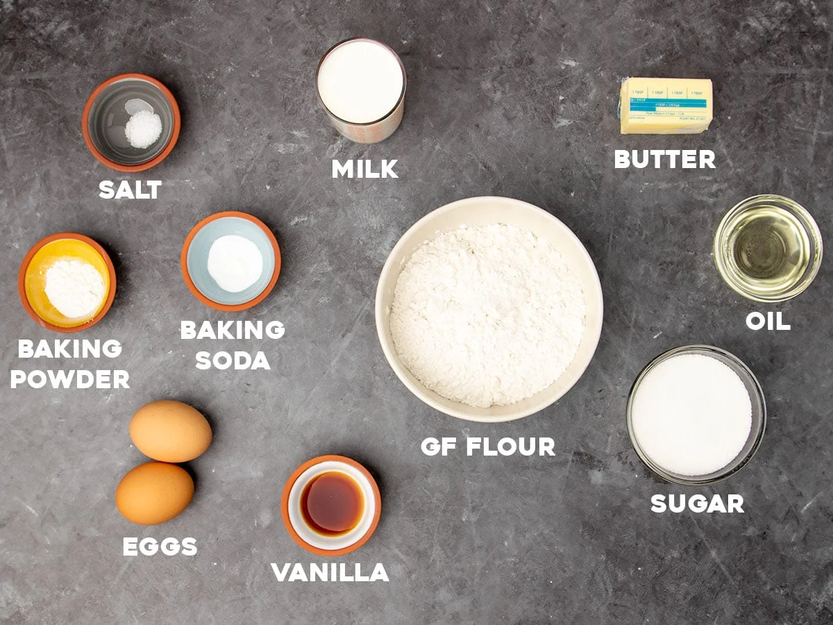 overhead image on black marbled surface with ingredients for gluten free strawberry shortcake cake in small bowls with white block letters naming each ingredient