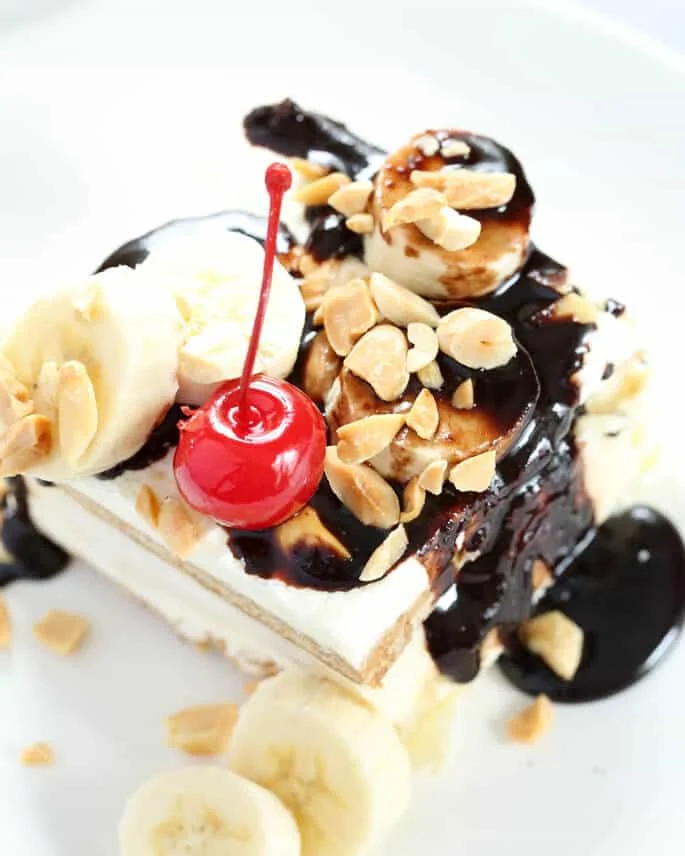 Overhead view of a banana split cake with a cherry, chocolate sauce, bananas and nuts on a white plate