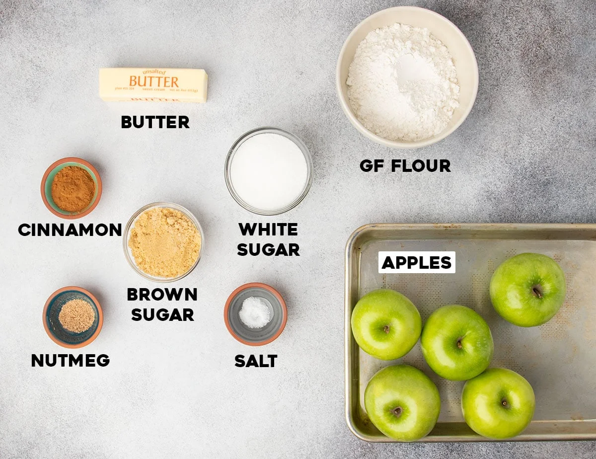 Overhead view of apples, gluten free flour, sugars, butter, and spices arranged on a gray surface.