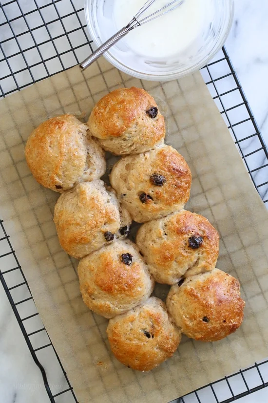 Overhead view of baked hot cross buns on a parchment-lined rack next to a bowl of glaze with a whisk.