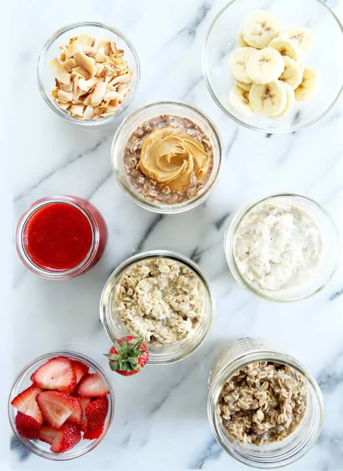 Overhead view of different jars of overnight oats and bowls of strawberries, strawberry puree, bananas and coconut chips