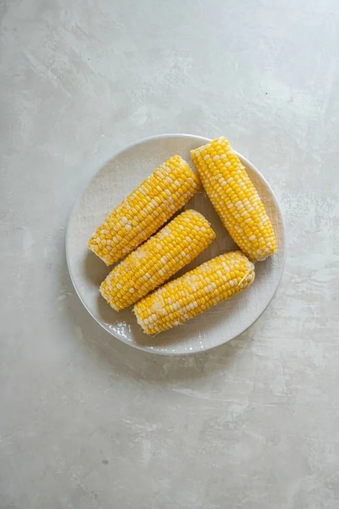 Overhead view of four unprepared ears of corn on a white plate.
