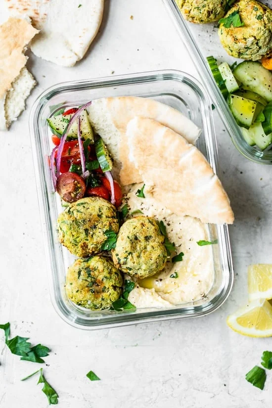 Overhead view of homemade falafel in meal prep container with pita bread slices, hummus, and a tomato cucumber salad.