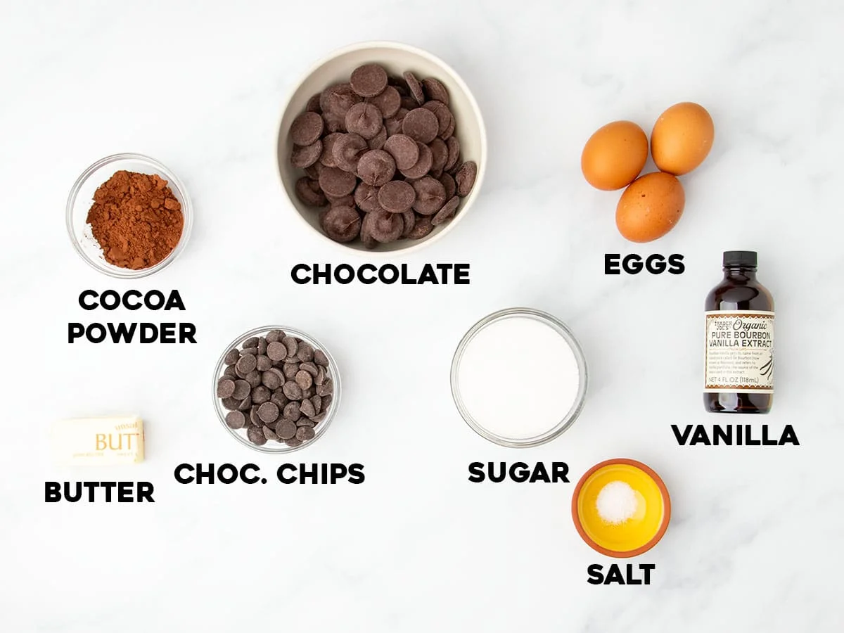 Overhead view of labeled ingredients for flourless brownies, including chocolate, cocoa, eggs, sugar, and butter
