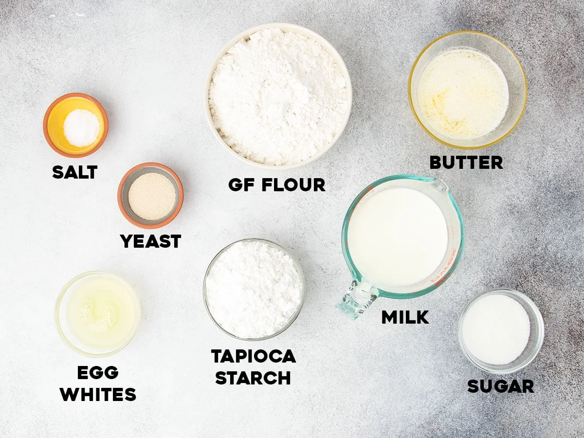 Overhead view of labeled ingredients required to make the recipe. Including gluten free flour, tapioca starch, yeast, salt, sugar, egg whites, milk, and melted butter.