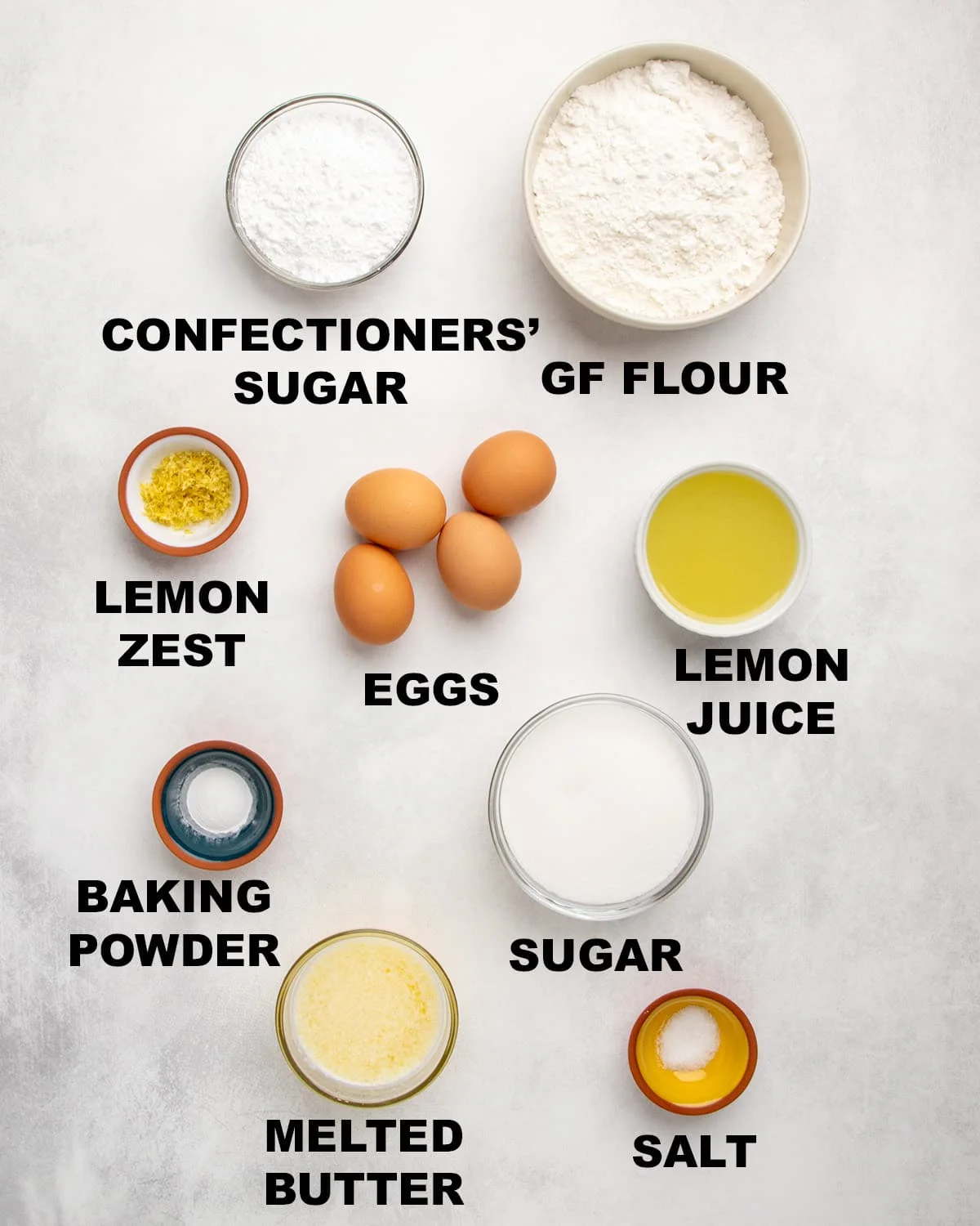 Overhead view of measured ingredients in bowls with labels including confectioners’ sugar, gluten free flour, lemon zest, eggs, lemon juice, baking powder, sugar, melted butter, and salt.