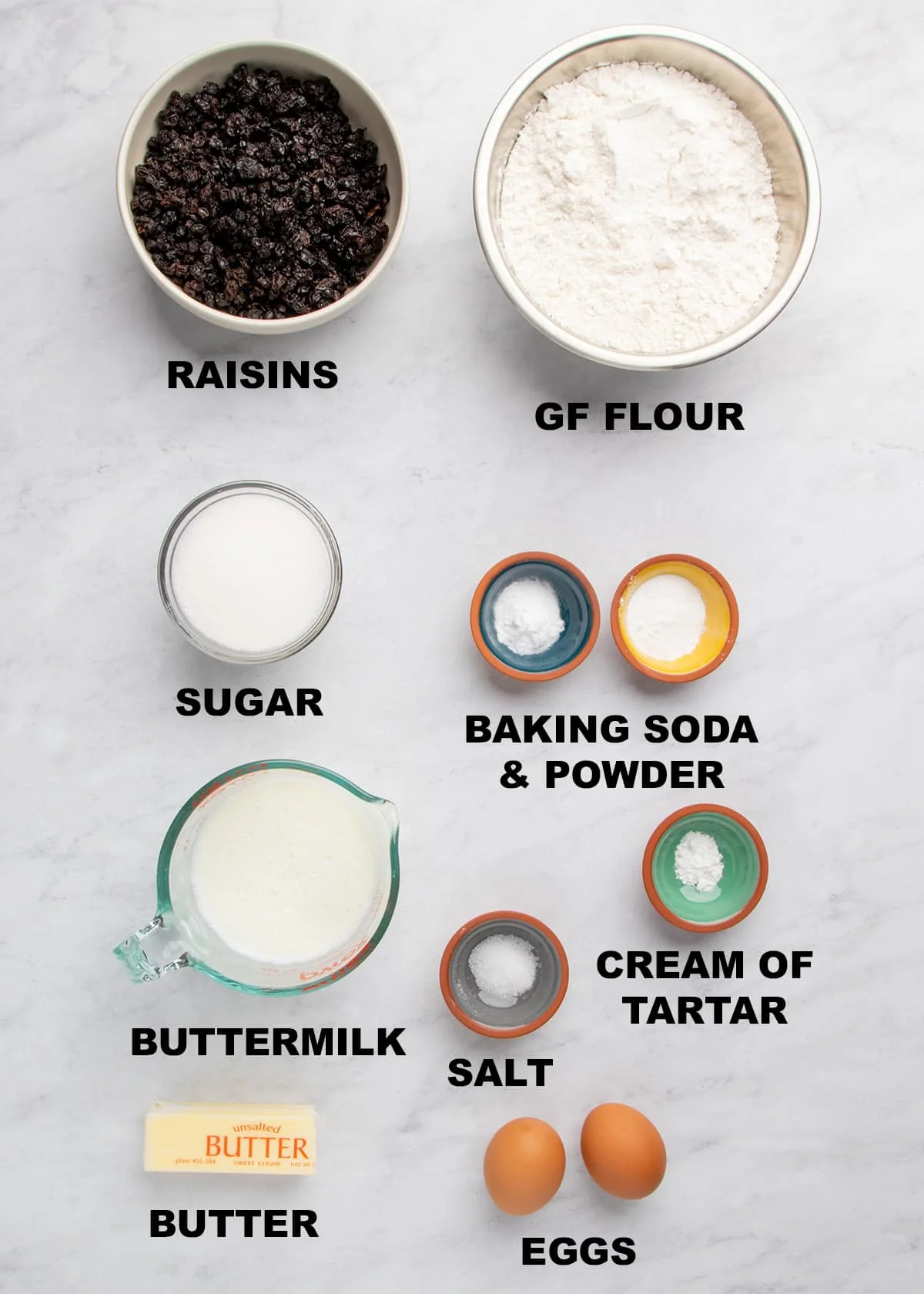 Overhead view of measured ingredients on a marble surface labeled raisins, gluten free flour, sugar, baking soda and baking powder, cream of tartar, salt, buttermilk, butter, and eggs.