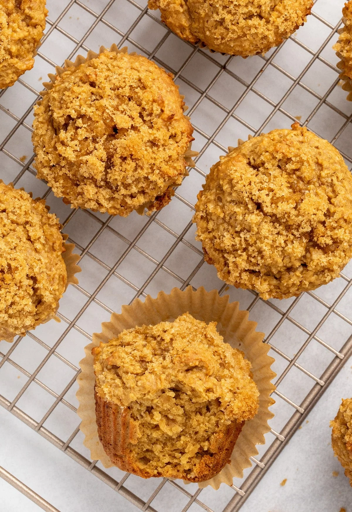 Overhead view of multiple baked muffins in paper liners on a wire cooling rack with one at the bottom of the view on its side with a bite taken.