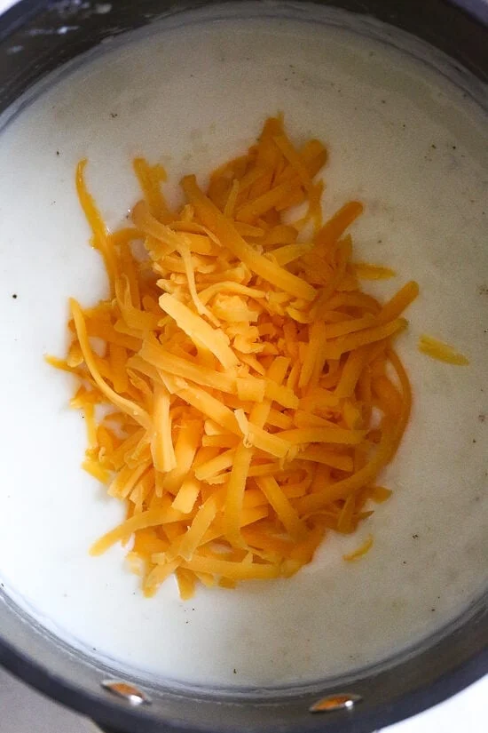 Overhead view of shredded cheddar cheese added to white sauce in a pot.